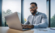 © Pixelowy_czarodziej - A smiling young man in a suit sits in the office at a desk and works on a laptop, typing on a keyboard, texting, working with data