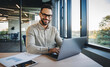 © Pixelowy_czarodziej - A smiling young man in a suit sits in the office at a desk and works on a laptop, typing on a keyboard, texting, working with data
