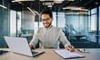 © Pixelowy_czarodziej - A smiling young man in a suit sits in the office at a desk and works on a laptop, typing on a keyboard, texting, working with data