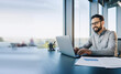 © Pixelowy_czarodziej - A smiling young man in a suit sits in the office at a desk and works on a laptop, typing on a keyboard, texting, working with data