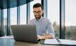 © Pixelowy_czarodziej - A smiling young man in a suit sits in the office at a desk and works on a laptop, typing on a keyboard, texting, working with data