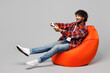 © ViDi Studio - Full body young happy Indian man he wears red shirt casual clothes sit in bag chair hold in hand play pc game with joystick console isolated on plain grey color background studio. Lifestyle concept.