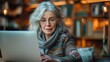 © visoot - Mature European woman, engaged in an online course on a laptop at a cozy home office, shelves with books in the background, warm ambient lighting. Generative AI.