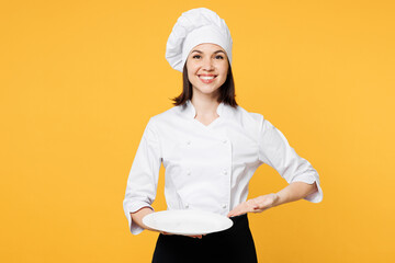 Wall Mural - Young fun chef cook baker woman wear white shirt black apron uniform toque chefs hat hold in hand point arm on empty plate look camera isolated on plain yellow background studio. Cooking food concept.