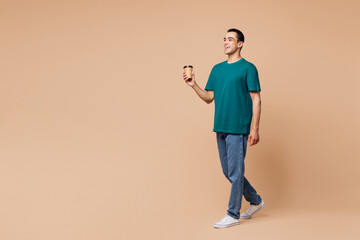 Full body fun young middle eastern man wear blue t-shirt casual clothes hold takeaway delivery craft paper brown cup coffee to go isolated on plain pastel beige background studio. Lifestyle concept.