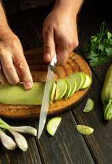 Before stewing, the zucchini must be cut. A man hands with a knife cut ripe zucchini on a wooden board. Zucchini diet at home.