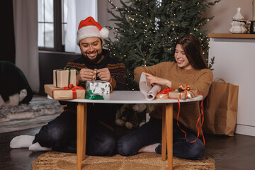 positive young millennials floor Christmas tree wrapping presents together. man wearing Santa hat. concept: gift wrapping for christmas on december 25. winter weekend holidays with the family 