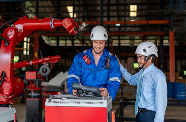 young caucasian male in safety gears holding remote control a robot arm welding machine talking with an old experienced manager to demonstrate robot readiness for operation in industrial factory