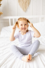 a little cute girl child on a white cotton bed at home is fooling around playing with tails and smiling, the child is playing and messing around at home in the morning