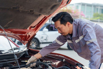 Asian male mechanic in gray uniform inspecting engine under open hood of red car outdoors. Focused on repair work, showing concentration and skill. Represents professionalism, technical expertise,