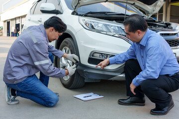 Wall Mural - Asian male insurance officer in uniform crouching by vehicle and using smartphone to document bumper damage while client points at specific area. Emphasizes detailed inspection, customer communication