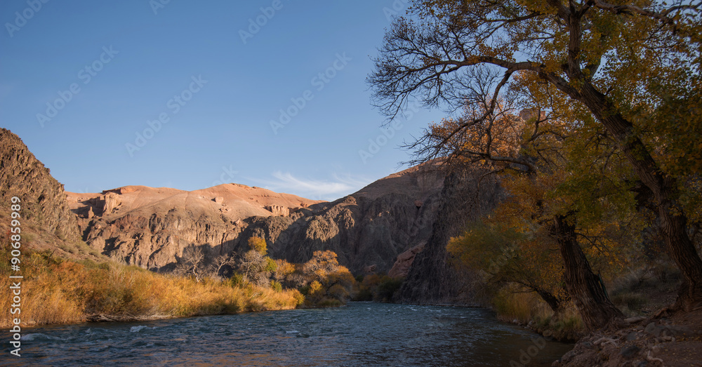Amber foliage frames a mountains river, meandering at the foot of towering, rugged cliffs under the gentle embrace of a clear azure sky.