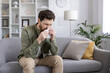 © Liubomir - Man sneezing into tissue while sitting on the couch. Indoor scene with man having cold or allergies, showing discomfort and illness.