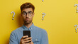 © Carmen - Young Caucasian man with confused expression while holding smartphone, studio shot against yellow background and white question marks