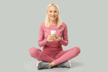 Poster - Mature woman in sportswear with cup of coffee sitting on grey background