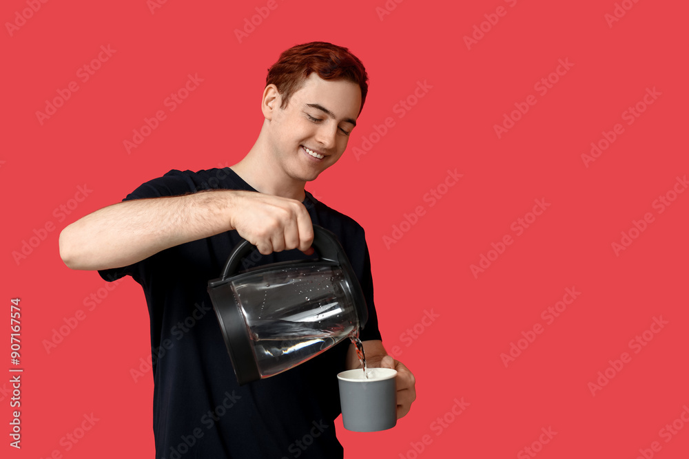 Handsome young man pouring hot water from modern electric kettle into mug on red background