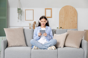 Poster - Beautiful young Asian woman in headphones listening to audiobook with modern tablet computer and sitting on sofa in living room