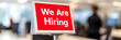 © Antto-AI - A red 'We Are Hiring' sign displayed prominently in a modern office setting, with blurred background of the workplace.
