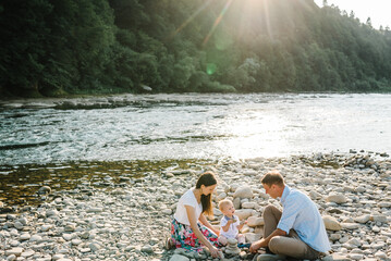 Mother, father, baby son sitting and playing on stones on beach near lake. Kid play with parents at sunset closeup. Happy family holiday on summer day. Spending time together in mountains.