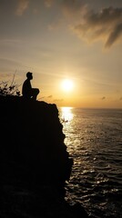 Poster - Silhouette of a man sitting on a cliff overlooking the ocean at sunset.