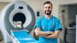 © Felippe Lopes - Smiling male doctor in blue scrubs stands in a medical office with an MRI machine in the background.