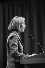 Wall Mural - A woman stands behind a podium with a microphone. She is wearing a gray jacket and she is giving a speech