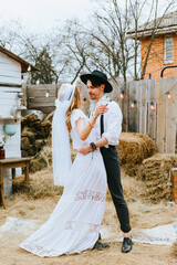 Wall Mural - wedding boho style ceremony young couple newlyweds bride in white dress with veil and groom in hat and jeans with suspenders hugging and having fun at barn of country house, shot in motion