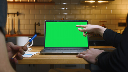 Two anonymous men working with laptop computer with green mock up screen on table in the kitchen pointing screen with hand. Co-workers working home office, remote work, technology concept	
