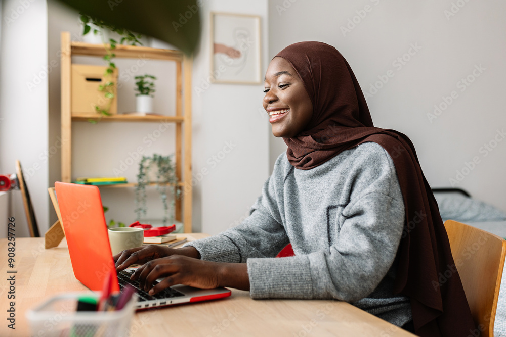 Happy black female student in muslim headscarf using laptop at home ...