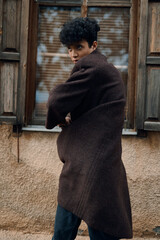 Wall Mural - Fashionable young man in a brown coat leaning against a window with a tilted head and hands on hips