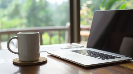 Wall Mural - A white mug and a laptop on a wooden table in front of a window with a view of trees.