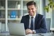© Philipp - A confident businessman in a suit smiling while working on a laptop in a contemporary office environment, reflecting productivity and professionalism.