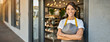 © Olga Gorkun - Entrepreneur female shop owner stands confidently with her arms crossed, wearing a gray apron and smiling in front of her store, which has a cozy, inviting atmosphere.