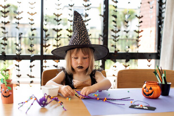 Wall Mural - A cute girl in a witch costume makes a funny spider for Halloween with her own hands. Children's crafts for Halloween