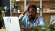 © VK Studio - A man in glasses sits at a desk, hand on his head, looking frustrated while staring at a laptop, surrounded by plants and shelves.