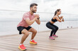 © muse studio - A man and woman perform squats together on a wooden deck by the water, focusing on their workout.