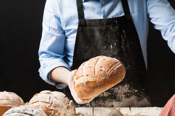 Male baker wearing black apron holding a freshly baked rustic bread splashing flour in front of a white wooden table filled with different kinds of loaves and jutte fabric