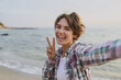© ViDi Studio - Close up young happy woman wear shirt casual clothes do selfie shot pov on mobile cell phone show v-sign rest on sea ocean sand shore beach outdoor seaside in summer day free time. Lifestyle concept.