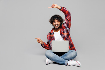 Poster - Full body young IT Indian man he wears red shirt casual clothes sits hold use work on laptop pc computer point finger aside on area isolated on plain grey color background studio. Lifestyle concept.