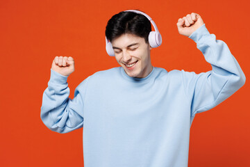 Poster - Young smiling happy fun man he wears blue sweatshirt casual clothes listen to music in headphones raise up hands dance isolated on plain red orange color background studio portrait. Lifestyle concept.