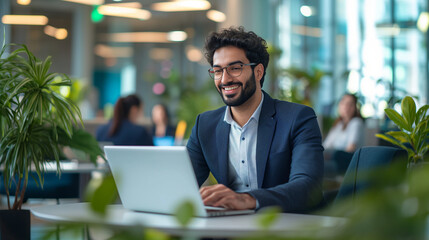 a happy middle eastern man in business attire working on his laptop at an open plan office with othe