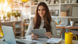 © @ArtUmbre - A woman with long brown hair, seated at a table in a brightly lit room. She is holding a tablet and appears to be smiling or engaged in something on the screen. Beside her is a laptop, a yellow mug