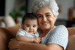 © Carmen - Senior Hispanic grandmother holding her grandchild, while sitting on cozy sofa, copy space