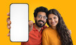 © Prostock-studio - A young Indian couple, both with dark hair and smiles, are posing against a yellow background. They are holding a smartphone with a blank screen, which is positioned in front of the camera.