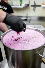 Poster - close-up of a gloved hand stirring a large metal pot filled with pink creamy soup, sprinkled with herbs, in an industrial kitchen setting. background is blurred, emphasizing the cooking process.