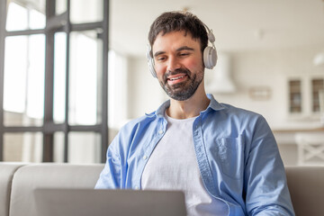 Poster - A man sits on a white couch in a living room with a window behind him. He is wearing headphones and smiling as he looks at the screen of a laptop.
