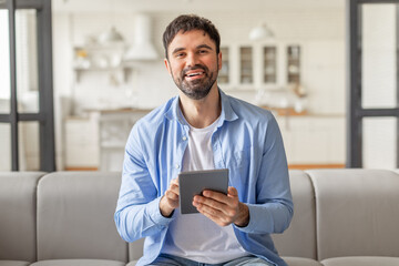 Poster - A man sits on a gray couch in his home and smiles while holding a tablet computer in his hands, websurfing or scrolling