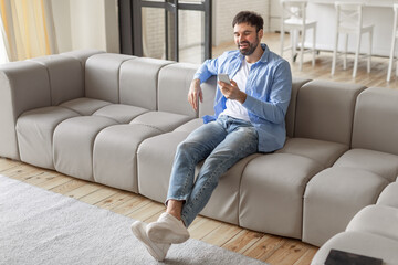 Poster - A man, dressed in jeans and a light blue shirt, sits on a light grey sofa in a modern living room. He is smiling as he uses his smartphone, copy space