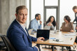 © Prostock-studio - A young businessman in a blue suit is working at a desk in a modern office. He is looking directly at the camera with a confident expression. He is sitting at a desk with a laptop computer