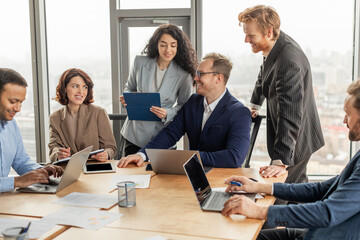 Poster - A group of diverse professionals, including a woman holding a clipboard, a man leaning against a chair, and a man typing on a laptop, are collaborating in a modern office setting